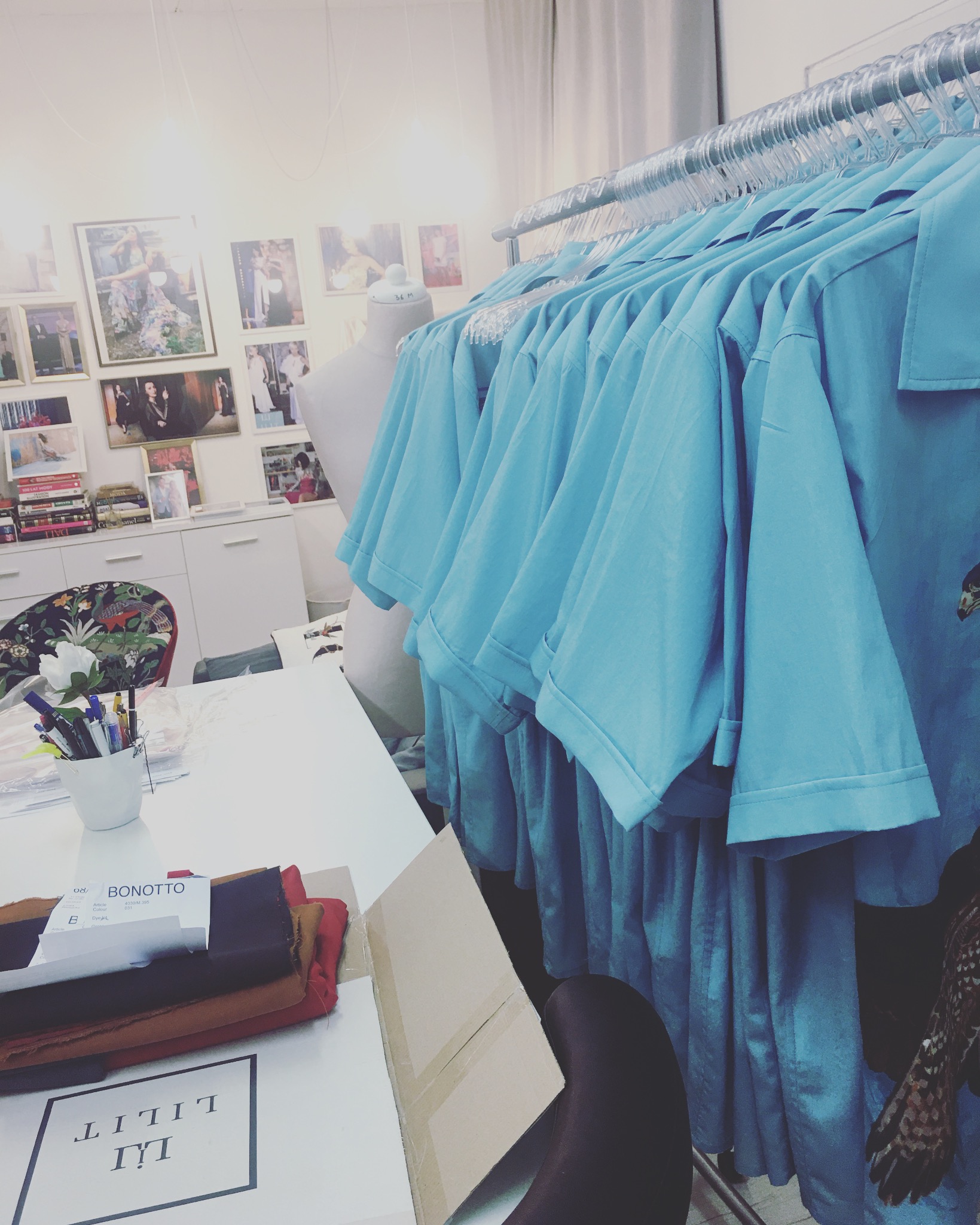 Atelier krawieckie: Rząd błękitnych koszul na wieszakach, manekin krawiecki w tle, stół z próbkami materiałów, kartonem i przyborami do szycia, zdjęcia projektów na ścianie.