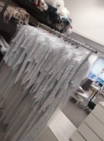 Rząd ubrań damskich z wzorem zebry, wiszących na wieszakach pod folią ochronną, w tle rolki materiałów w szwalni.