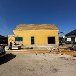 Royal Bud - Budowa domu szkieletowego w słoneczny dzień. Widoczne ściany z płyt OSB, rusztowanie i przygotowane materiały budowlane. Częściowo zadaszony dach. Dwóch pracowników w tle.