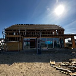 Royal Bud - Konstrukcja domu szkieletowego w trakcie budowy, widoczne rusztowania i elementy drewniane, jasne niebo w tle. Duże okna z niebieską folią ochronną.