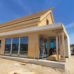 Royal Bud - Budowa domu szkieletowego: widok z zewnątrz, pracownicy montują elementy konstrukcyjne na jasnym, słonecznym tle. Widoczne duże okna i fragment dachu.