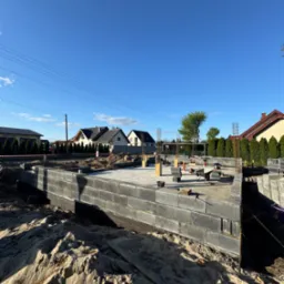 Fundamenty domu w trakcie budowy: bloczki betonowe, zbrojenie wystające ponad poziom fundamentu, piasek i narzędzia budowlane w otoczeniu. Słoneczny dzień.