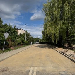 Royal Bud - Ułożona kostka brukowa na drodze dojazdowej, widok z perspektywy ulicy, zieleń po bokach, błękitne niebo z chmurami w tle. Widoczne białe pasy na asfalcie.