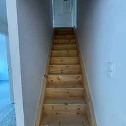 Royal Bud - Drewniane schody prowadzące na górę, z butelką środka czyszczącego na dole. Białe ściany i widoczne gniazdko elektryczne. Ujęcie z dołu, perspektywa w górę.