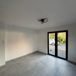 Royal Bud - Jasne wnętrze z szarymi płytkami podłogowymi i czarnymi drzwiami balkonowymi. Widok na zewnątrz z samochodem i zielenią. Minimalistyczna lampa sufitowa.