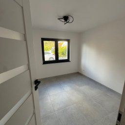 Royal Bud - Pusty pokój z szarymi płytkami podłogowymi, białymi ścianami i czarną ramą okienną. Widok przez otwarte drzwi na fragment pokoju i okno z widokiem na zewnątrz.