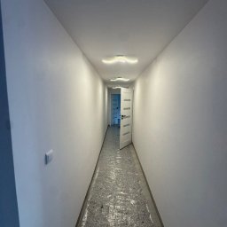 Royal Bud - Biały korytarz z otwartymi drzwiami i nowoczesnym oświetleniem sufitowym. Podłoga zabezpieczona folią ochronną. Świeżo pomalowane ściany.