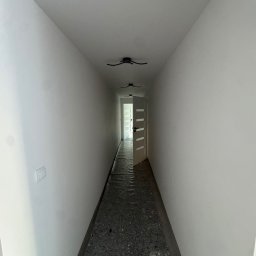 Royal Bud - Biały korytarz z otwartymi drzwiami na końcu, zabezpieczona podłoga, minimalistyczne lampy sufitowe. Widok w perspektywie z niskiego kąta.