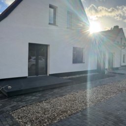 Royal Bud - Biały dom z ciemną stolarką okienną i drzwiami, widok z zewnątrz, z brukowanym podjazdem i kamiennym rabatem. Słońce tworzy efekt rozbłysku.