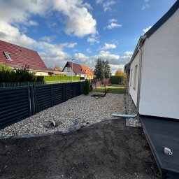 Royal Bud - Nowoczesne ogrodzenie panelowe w kolorze antracytu oddziela posesję. Część działki wysypana drobnymi kamieniami, przygotowany teren pod trawnik. Widok na domy w tle.