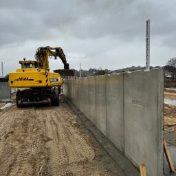 Royal Bud - Żółta koparka Atlas ustawia prefabrykowane betonowe panele ogrodzeniowe z wytłoczonymi numerami na placu budowy pod pochmurnym niebem.