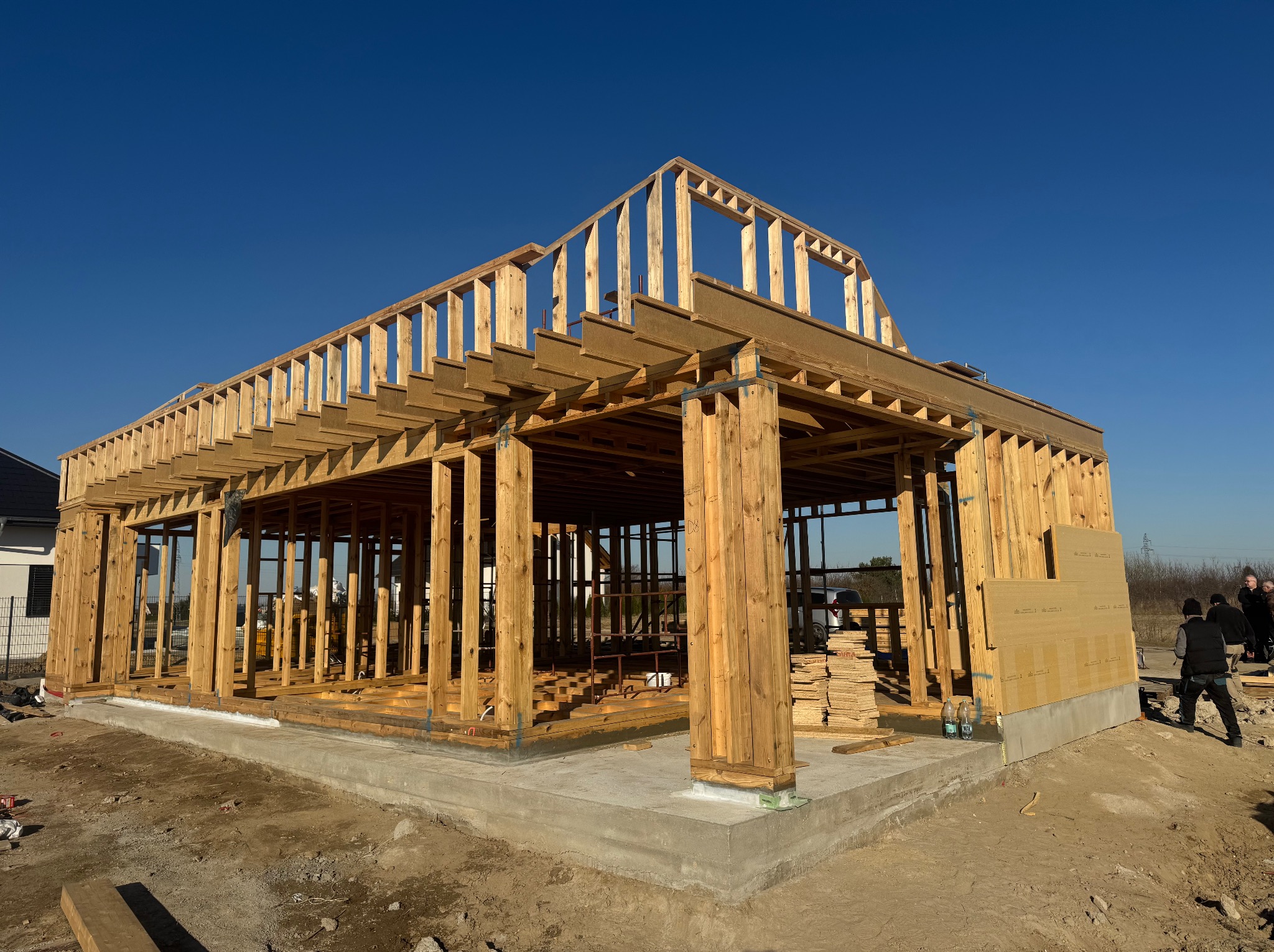 Konstrukcja szkieletowa domu w trakcie budowy, widoczne drewniane elementy nośne, fundament betonowy i stos płyt OSB na zewnątrz, błękitne niebo w tle.