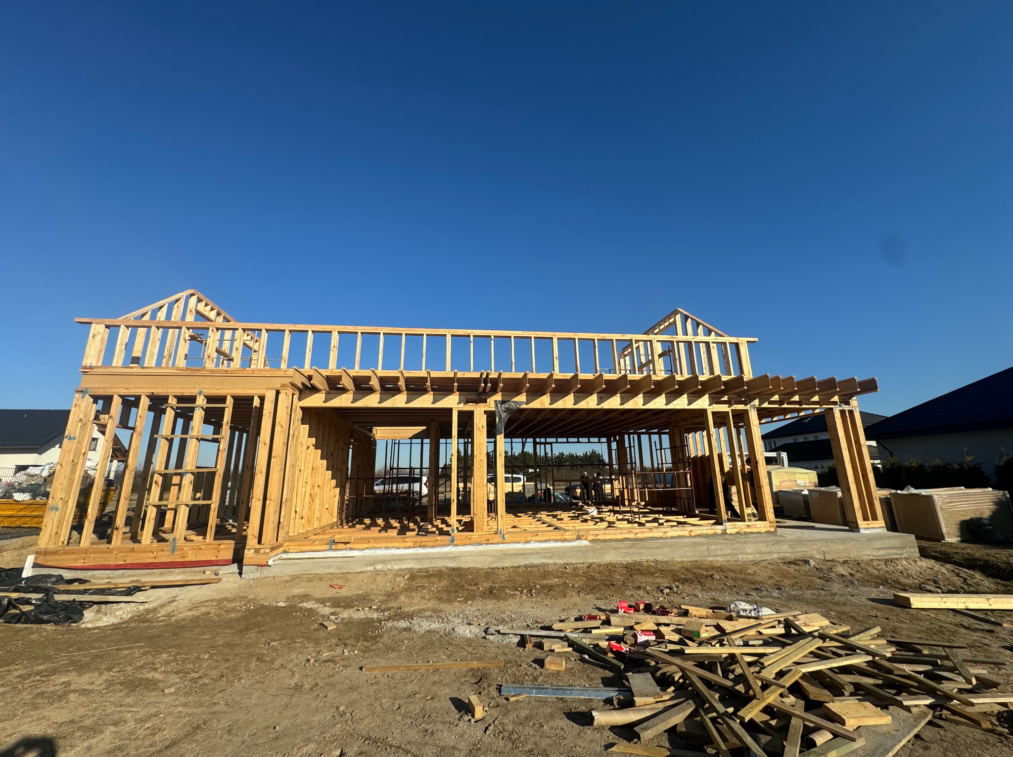 Wznoszenie konstrukcji domu szkieletowego na fundamencie, widoczne drewniane elementy konstrukcyjne, niebieskie niebo w tle, teren budowy z porozrzucanymi resztkami drewna.