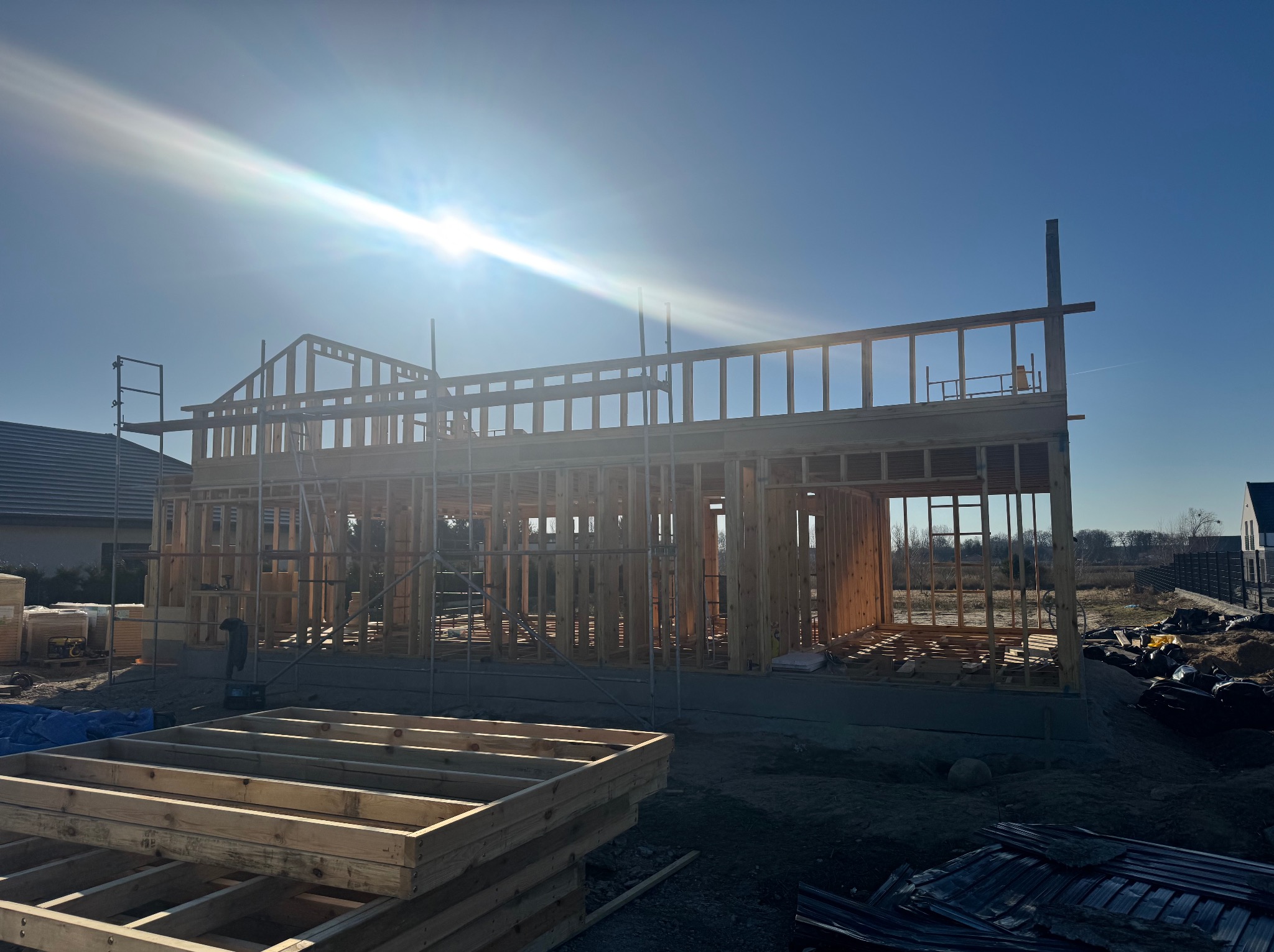 Konstrukcja domu szkieletowego w trakcie budowy, widoczne drewniane elementy nośne i szkielet ścian, w tle błękitne niebo i silne słońce.