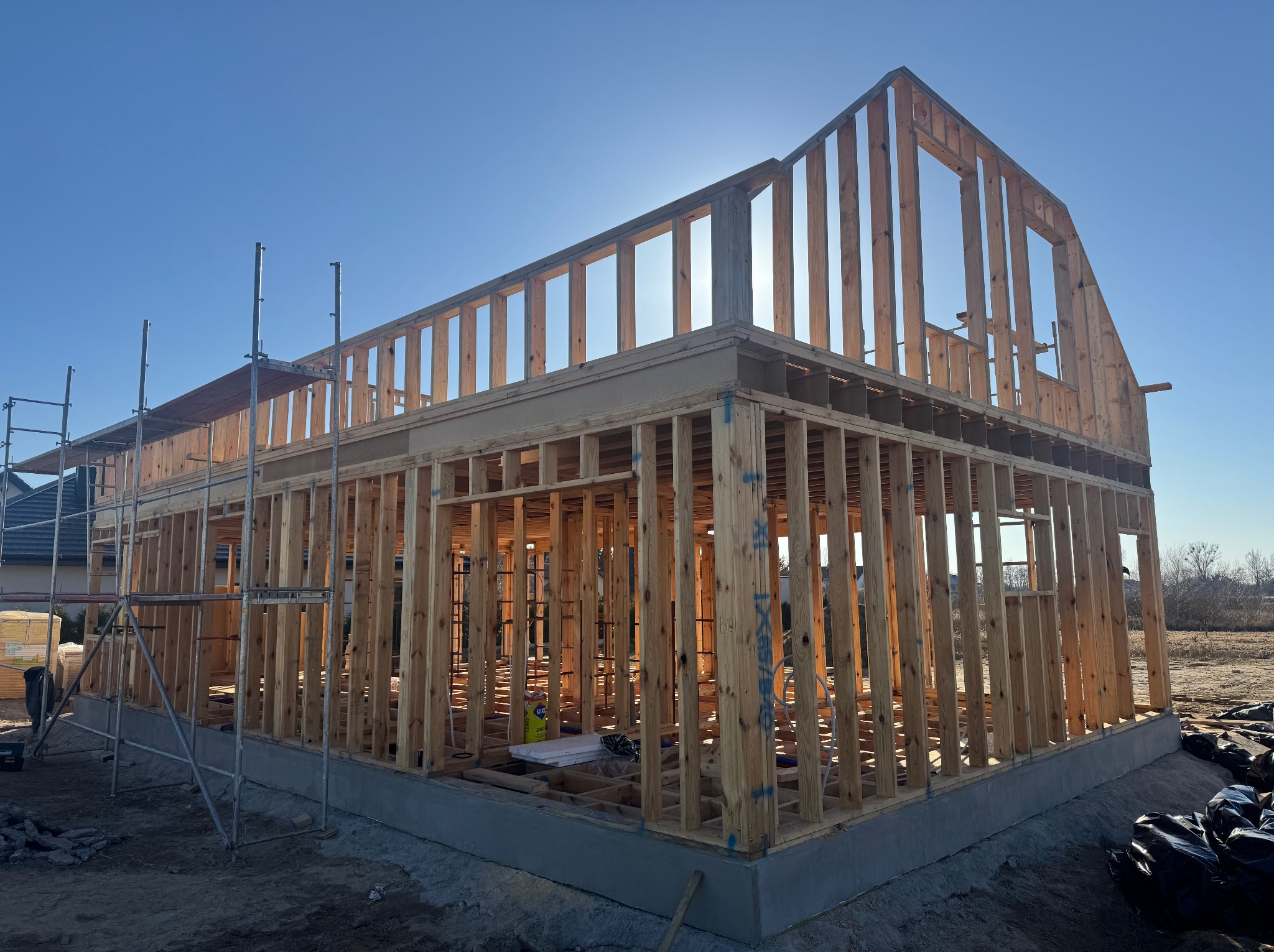 Surowy szkielet domu w trakcie budowy, widoczny na tle błękitnego nieba z silnym słońcem, z rusztowaniem po lewej stronie i materiałami budowlanymi składowanymi obok.