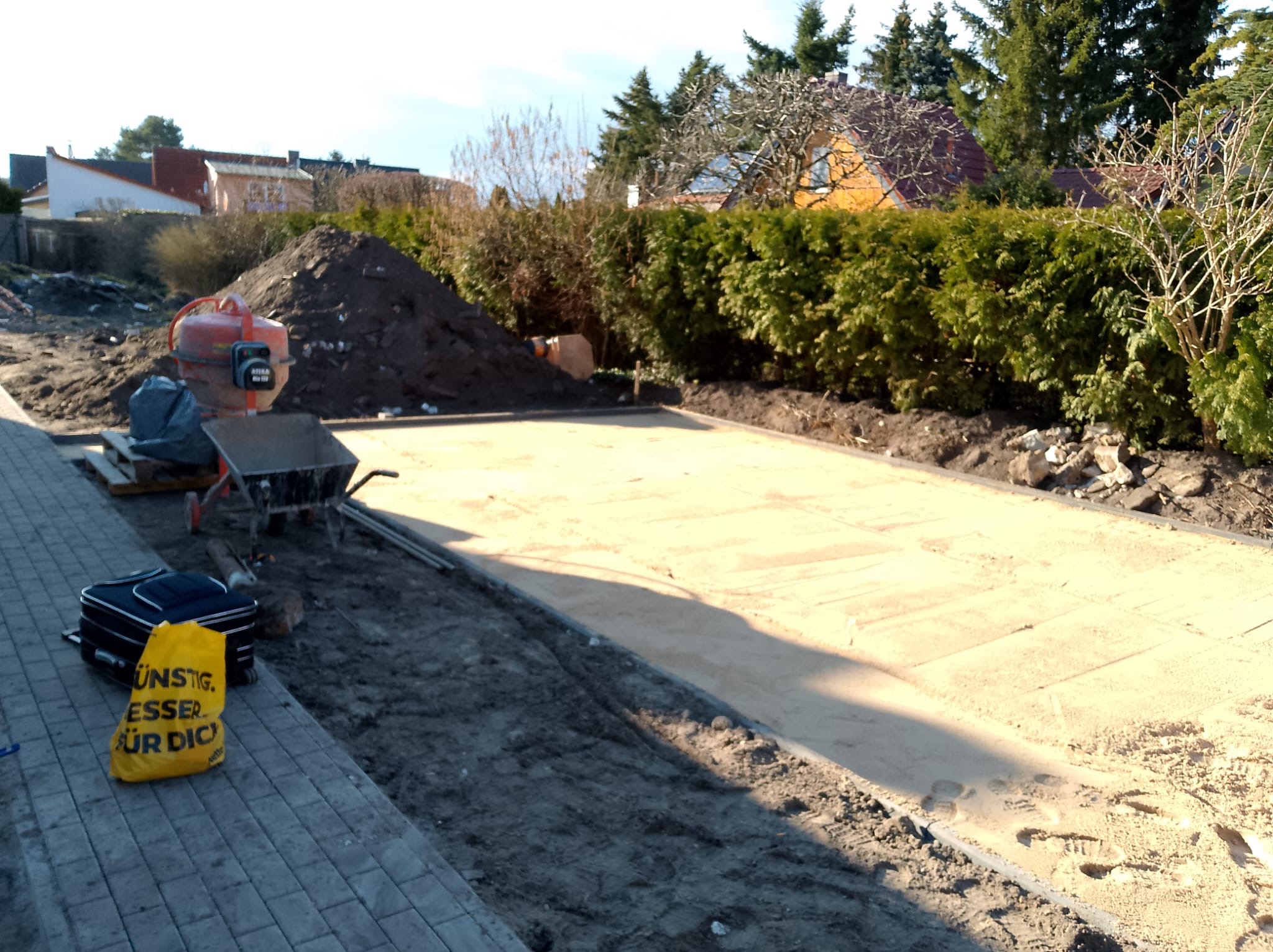 Przygotowanie podłoża pod kostkę brukową, widoczna warstwa piasku, obrzeża, betoniarka, taczka i narzędzia na placu budowy.