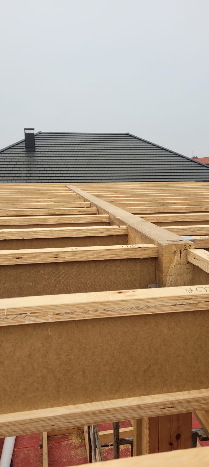 Konstrukcja drewniana dachu w trakcie budowy domu szkieletowego, widoczne elementy nośne i poszycie, w tle dach z czarnej dachówki i komin.