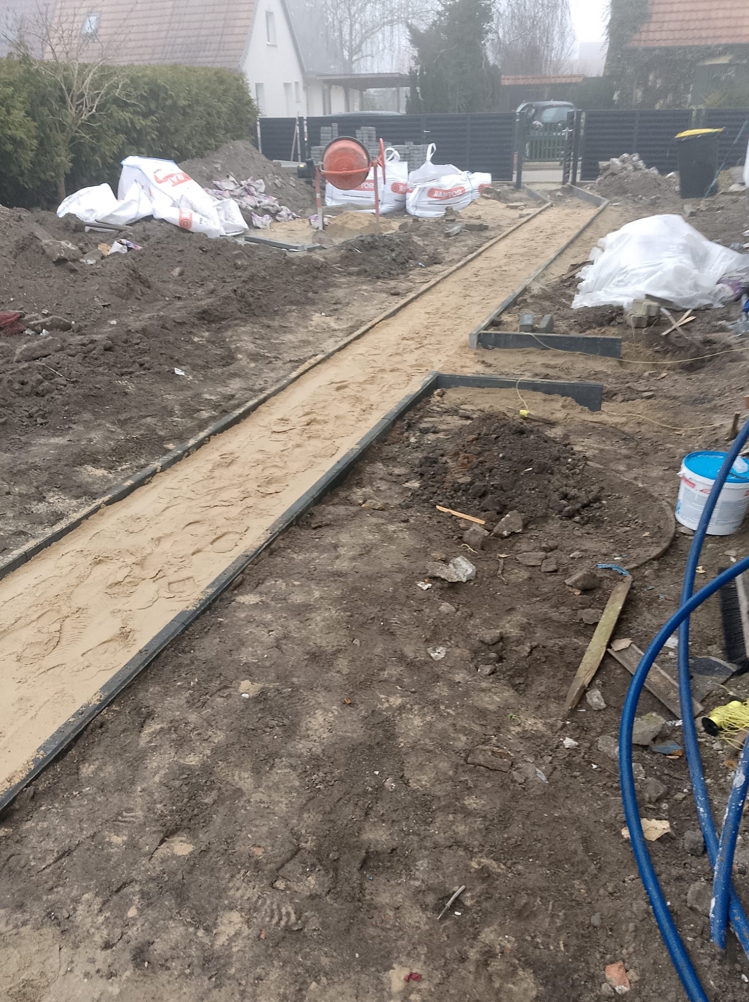 Przygotowanie podłoża pod ścieżkę ogrodową z obrzeżami, widoczne ślady stóp w piasku, w tle worki z materiałami budowlanymi, betoniarka i ogrodzenie.