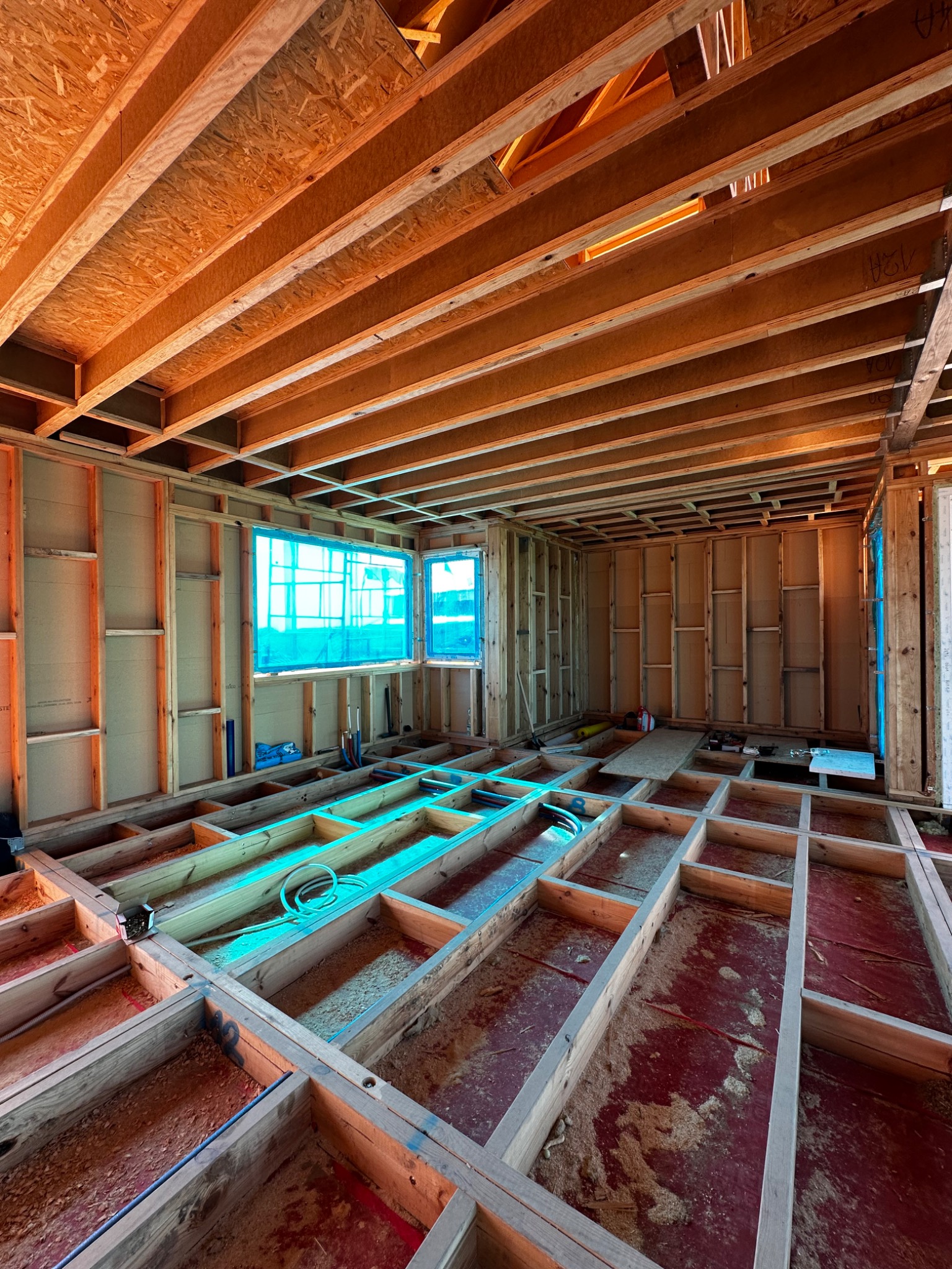 Wnętrze domu w trakcie budowy: drewniana konstrukcja szkieletowa ścian, belek stropowych i podłogi. Okna zabezpieczone folią. Widoczne instalacje.