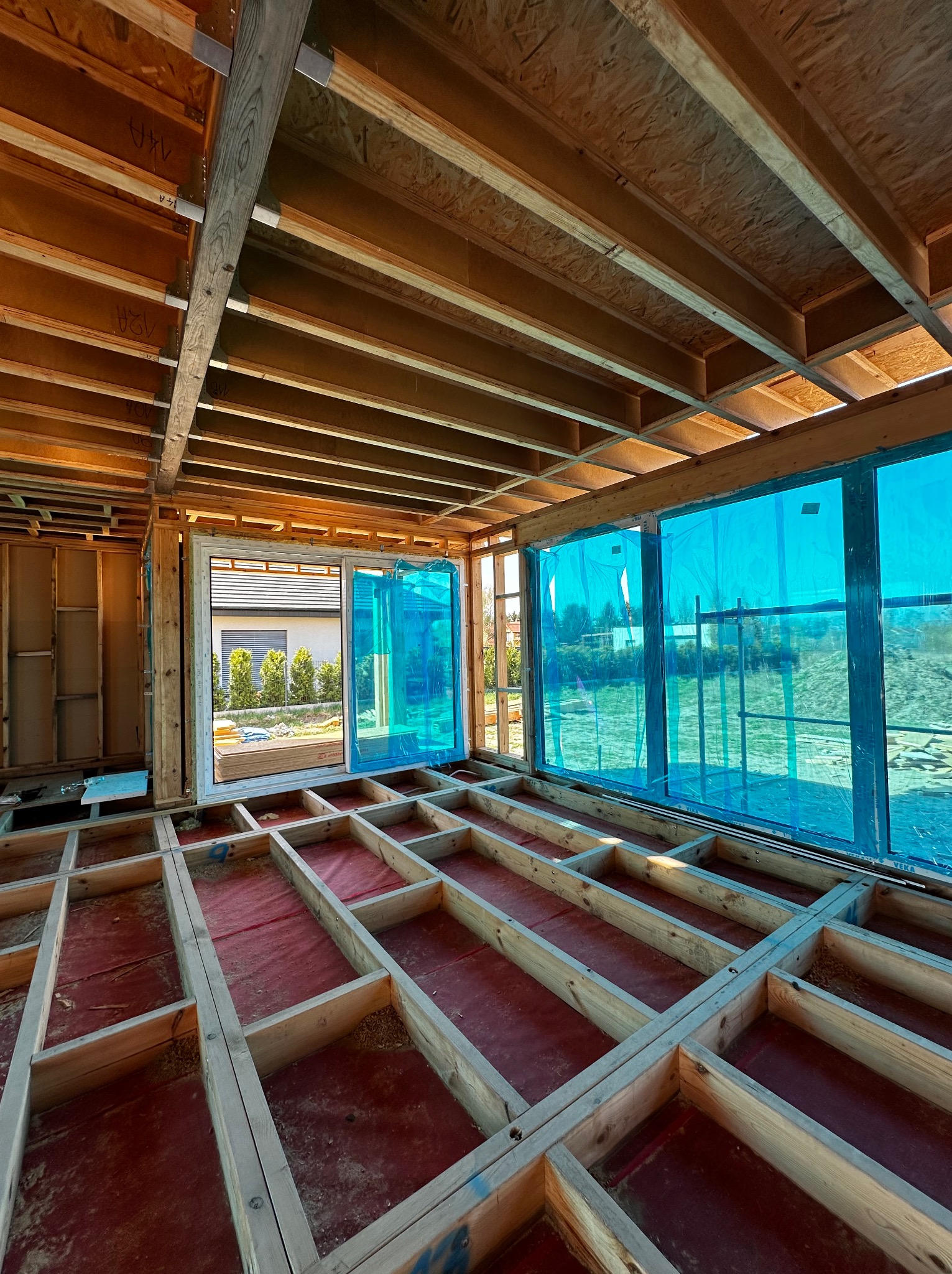 Wnętrze domu w trakcie budowy: drewniany szkielet podłogi na czerwonej folii, ściany z oknami zabezpieczonymi niebieską folią, widok na ogród. Etap budowy domu.