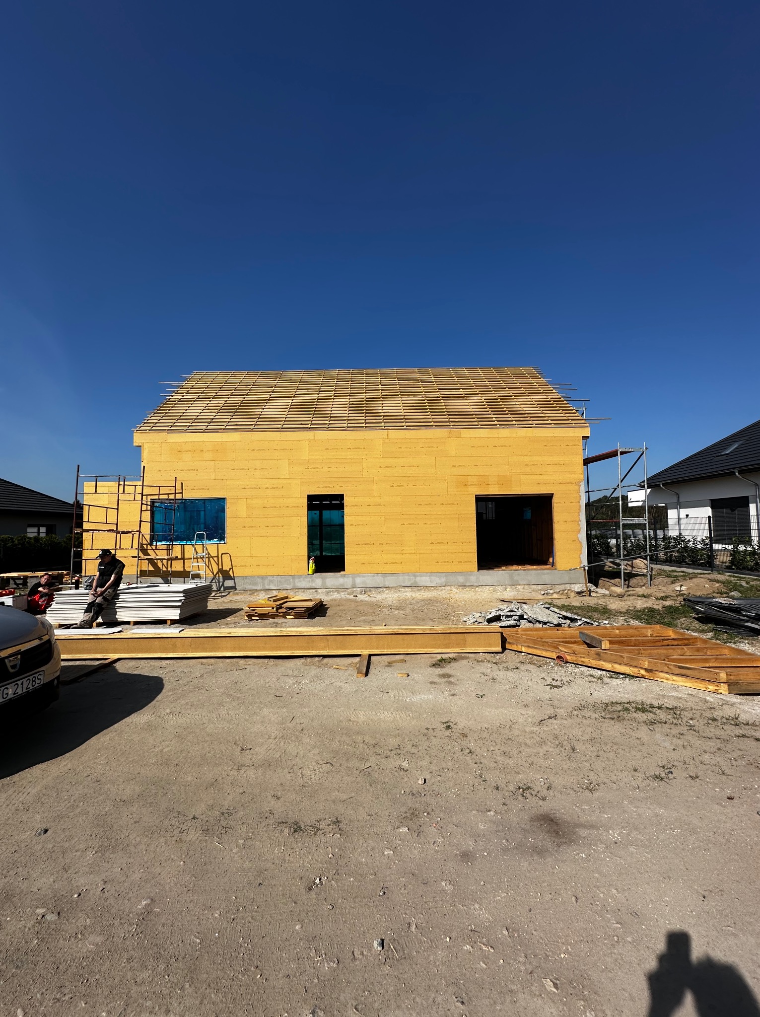 Budowa domu szkieletowego w słoneczny dzień. Widoczne ściany z płyt OSB, rusztowanie i przygotowane materiały budowlane. Częściowo zadaszony dach. Dwóch pracowników w tle.