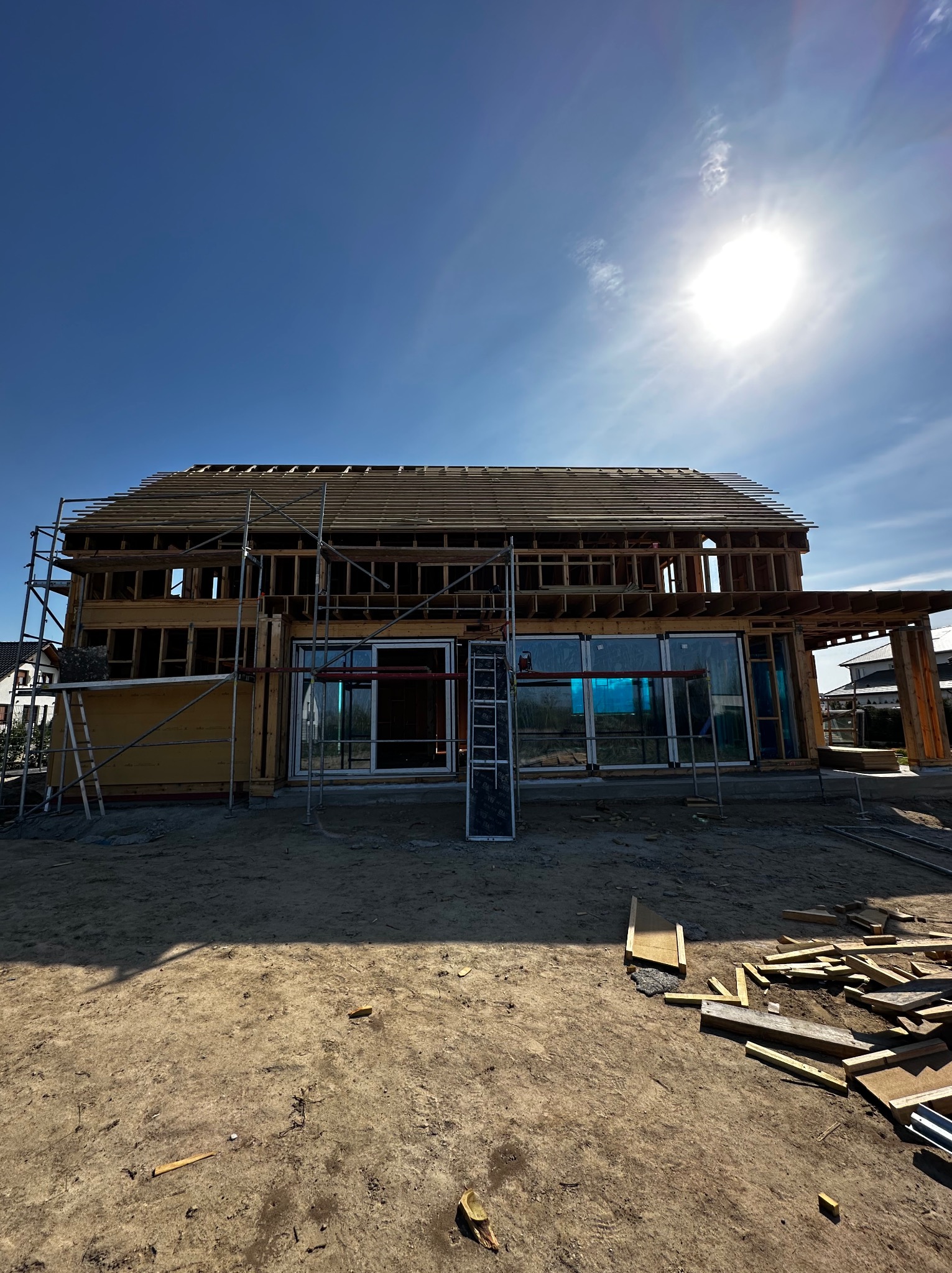 Konstrukcja domu szkieletowego w trakcie budowy, widoczne rusztowania i elementy drewniane, jasne niebo w tle. Duże okna z niebieską folią ochronną.