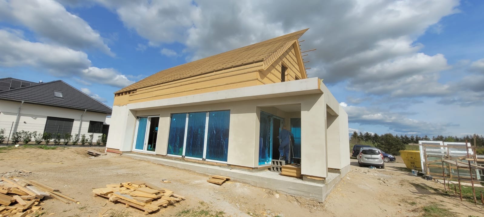 Niedokończony dom z drewnianym dachem i dużymi oknami zabezpieczonymi folią, w tle widoczny inny dom i błękitne niebo z chmurami.