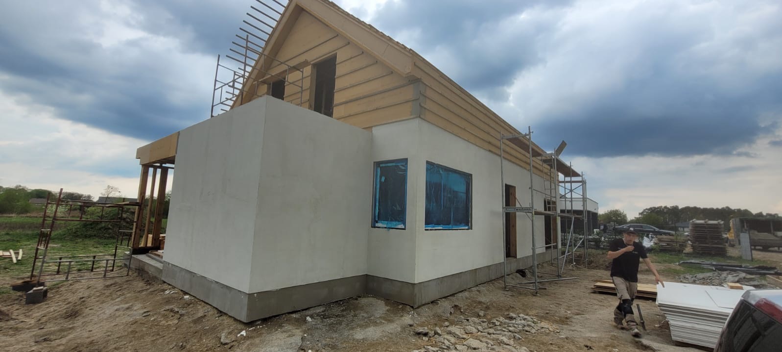 Budowa domu w trakcie realizacji: surowe ściany, drewniany szkielet dachu, okna zabezpieczone folią, rusztowania. W tle pochmurne niebo.