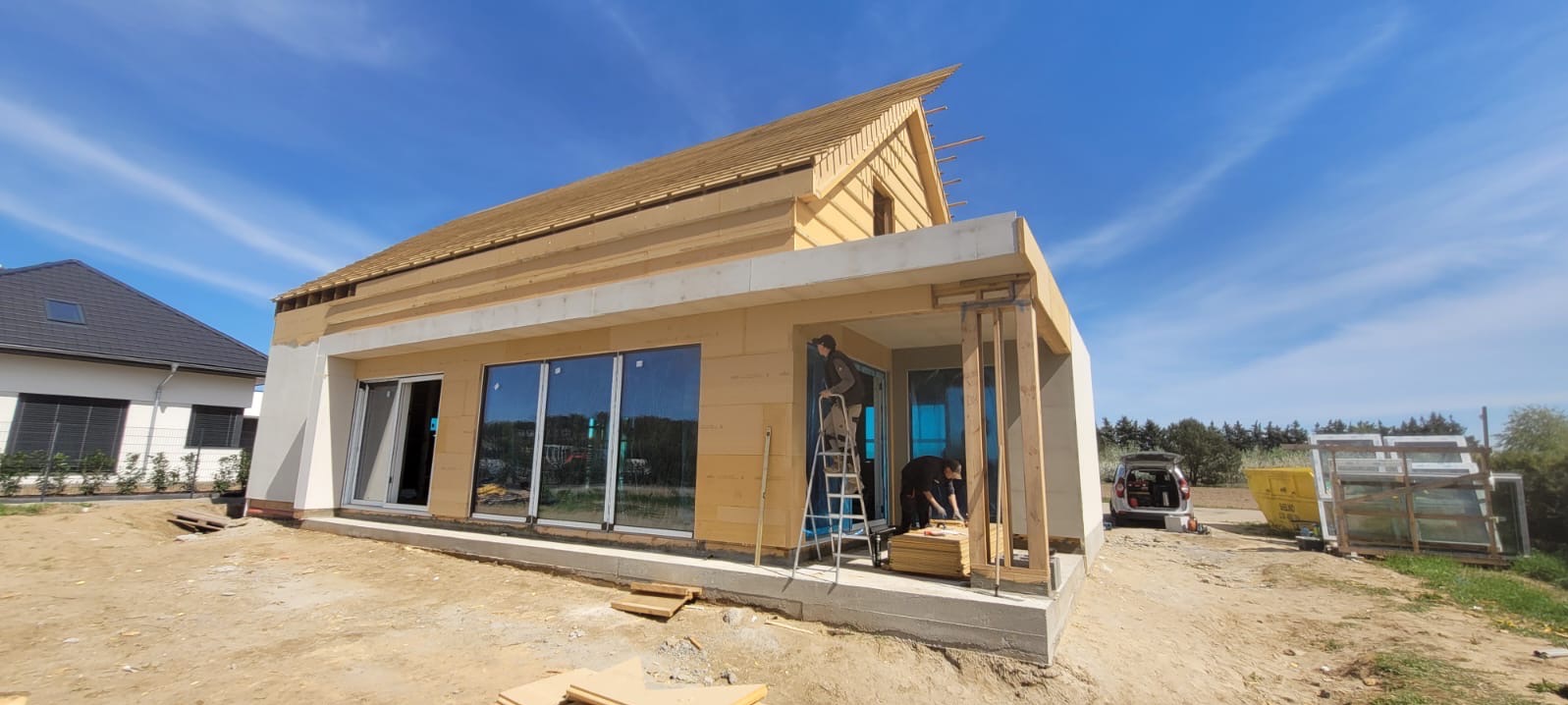 Budowa domu szkieletowego: widok z zewnątrz, pracownicy montują elementy konstrukcyjne na jasnym, słonecznym tle. Widoczne duże okna i fragment dachu.
