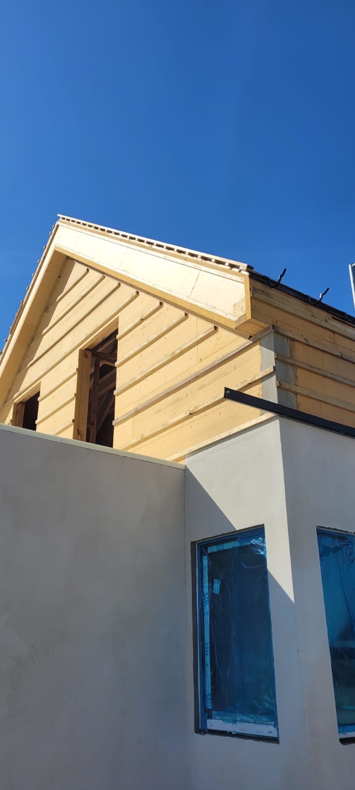 Budowa domu szkieletowego: widok na fragment ściany z oknami zabezpieczonymi folią i konstrukcją dachu w trakcie montażu, na tle błękitnego nieba.