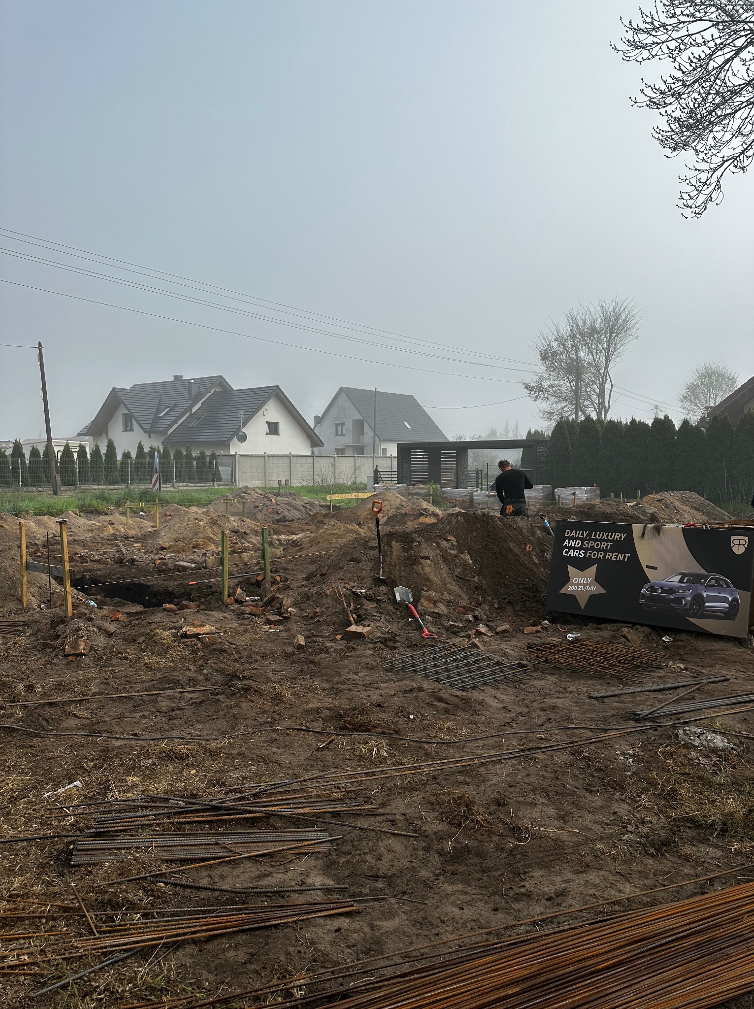 Przygotowanie terenu pod fundamenty domu: wykop, szalunki, zbrojenie, narzędzia. W tle zabudowa jednorodzinna i reklama wypożyczalni samochodów.