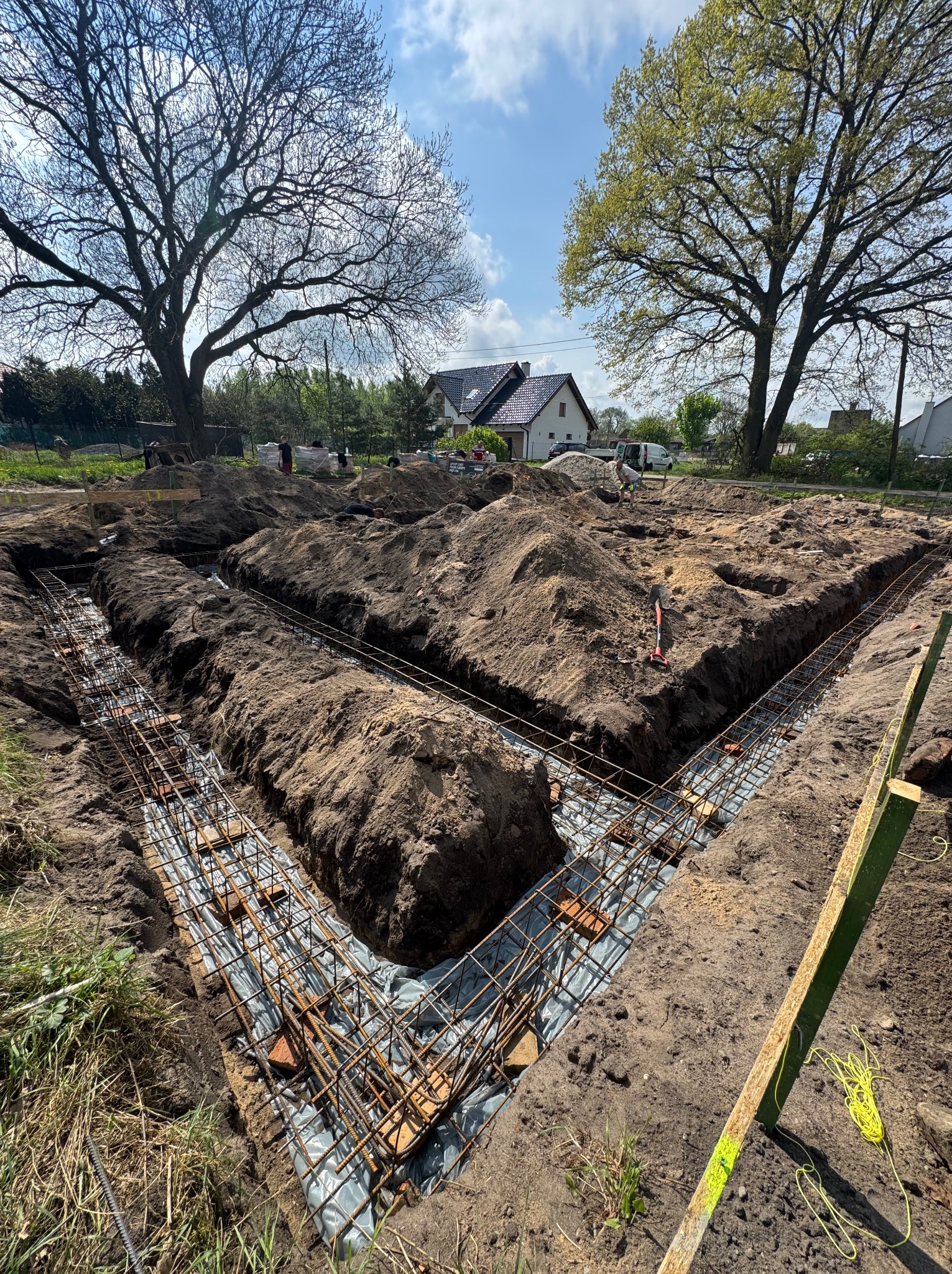 Przygotowanie zbrojenia fundamentów pod dom, widoczne wykopy, stalowe pręty zbrojeniowe i folia izolacyjna. W tle drzewa i budynek mieszkalny.