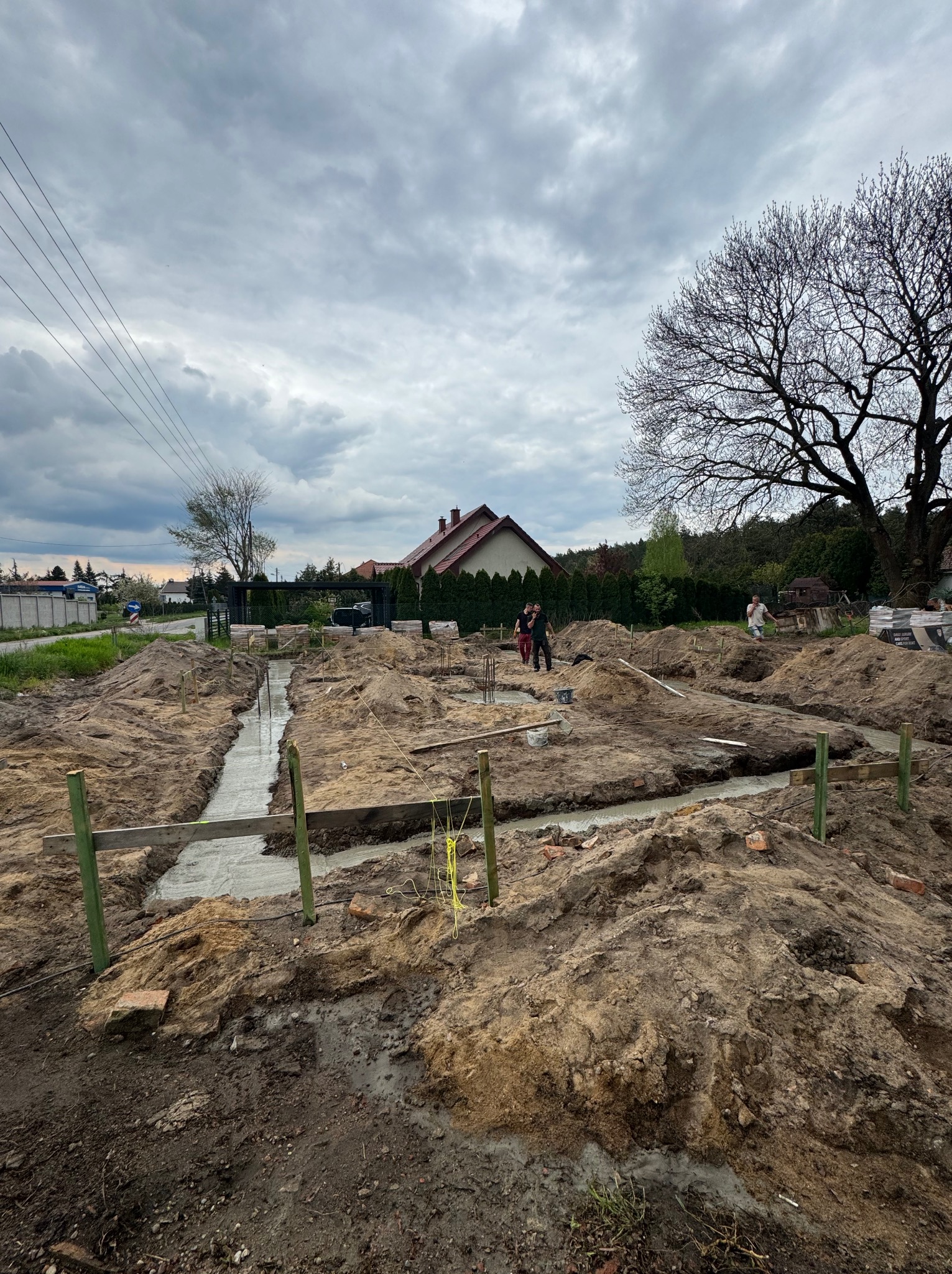 Zalane ławy fundamentowe z drewnianym szalunkiem, widoczne zbrojenia. W tle dom jednorodzinny i pochmurne niebo. Etap budowy domu.