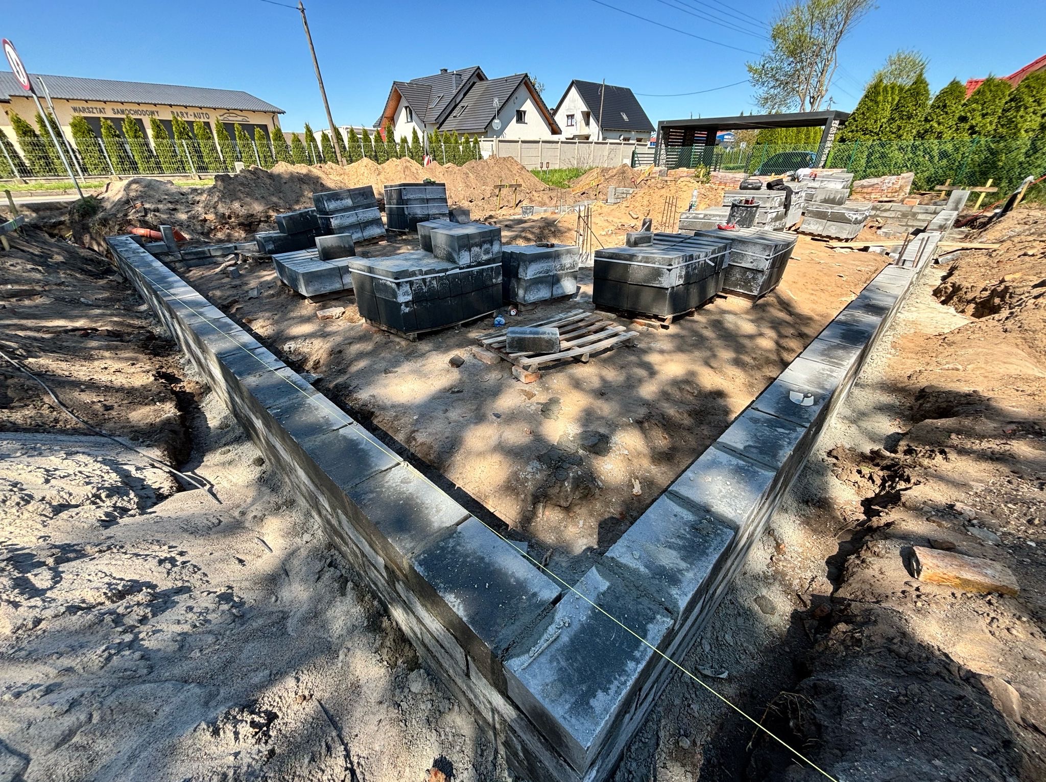 Fundamenty domu w trakcie budowy, widoczne bloczki betonowe i wykop, w tle domy jednorodzinne i błękitne niebo. Ujęcie z góry, podkreślające geometrię fundamentów.