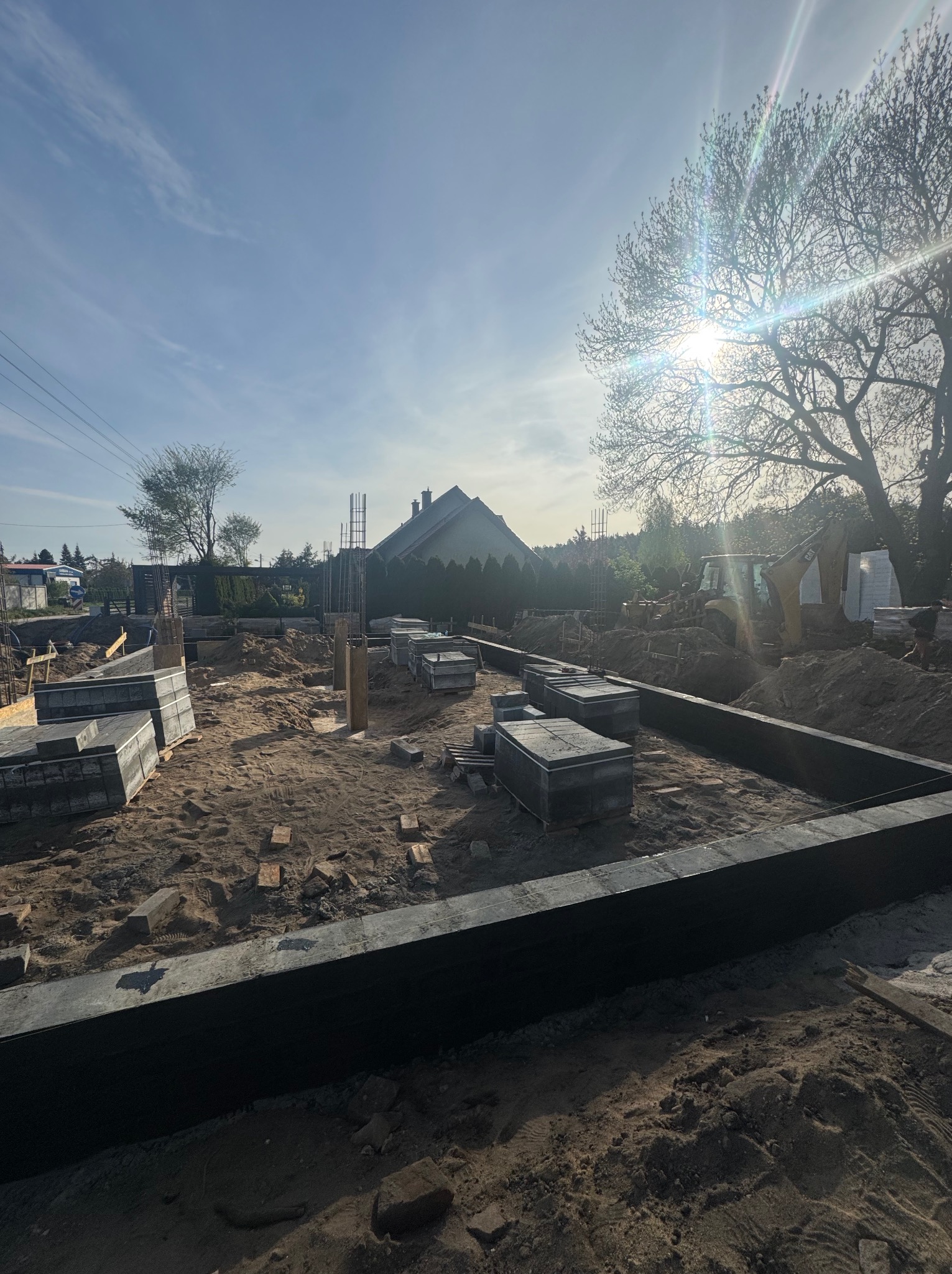 Fundamenty domu w trakcie budowy, z widocznym zbrojeniem i betonowymi elementami szalunkowymi, na tle błękitnego nieba z promieniami słońca.
