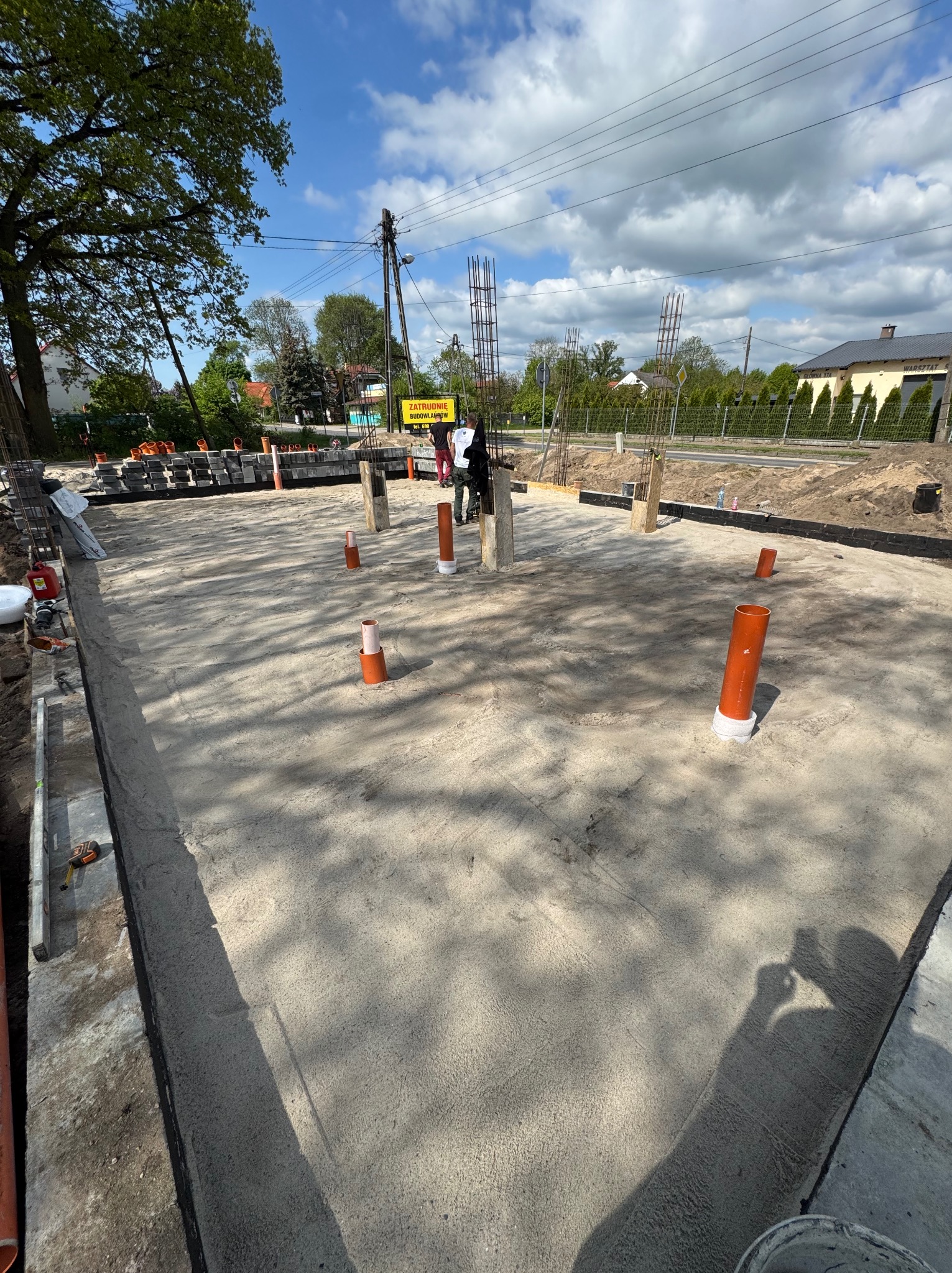 Zalana płyta fundamentowa z wystającymi pionowymi rurami kanalizacyjnymi i zbrojeniami w tle, przygotowana do dalszych prac budowlanych.