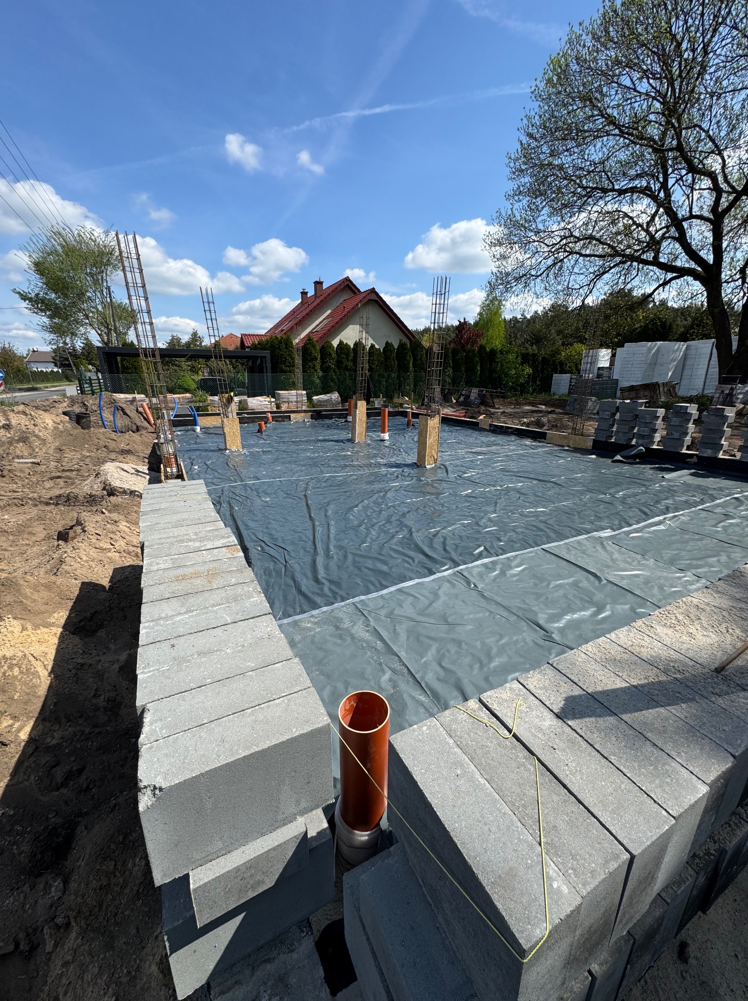 Fundamenty domu w trakcie budowy: szalunek z bloczków betonowych, folia hydroizolacyjna, wystające pręty zbrojeniowe, rury instalacyjne, błękitne niebo.