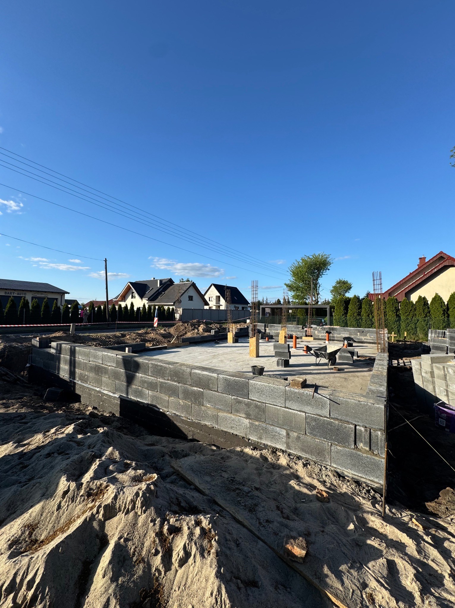 Fundamenty domu w trakcie budowy: bloczki betonowe, zbrojenie wystające ponad poziom fundamentu, piasek i narzędzia budowlane w otoczeniu. Słoneczny dzień.