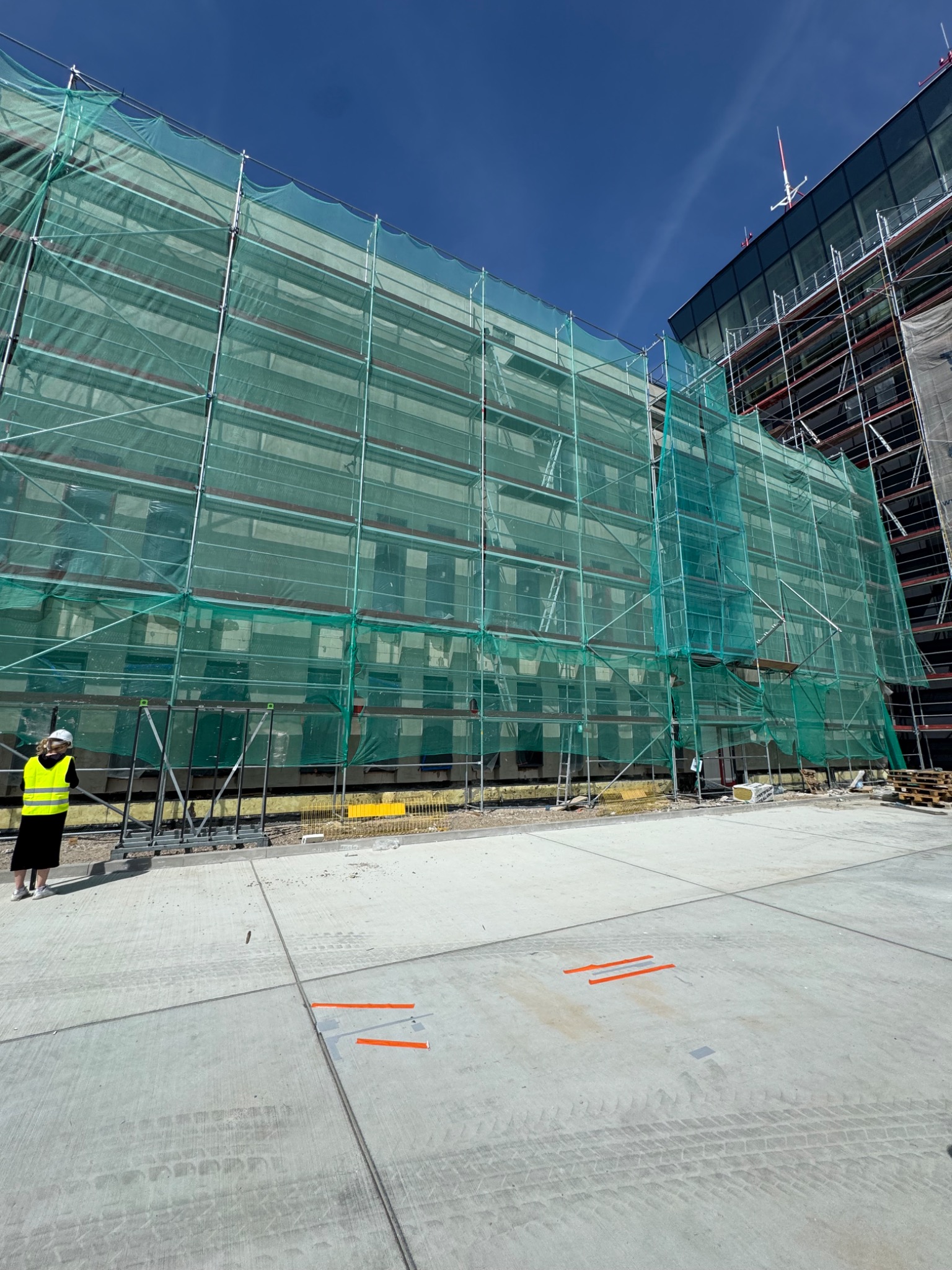 Budynek w trakcie ocieplania osłonięty siatką ochronną, widoczne rusztowania i pracownik w kamizelce. Ujęcie z dołu, betonowy plac.