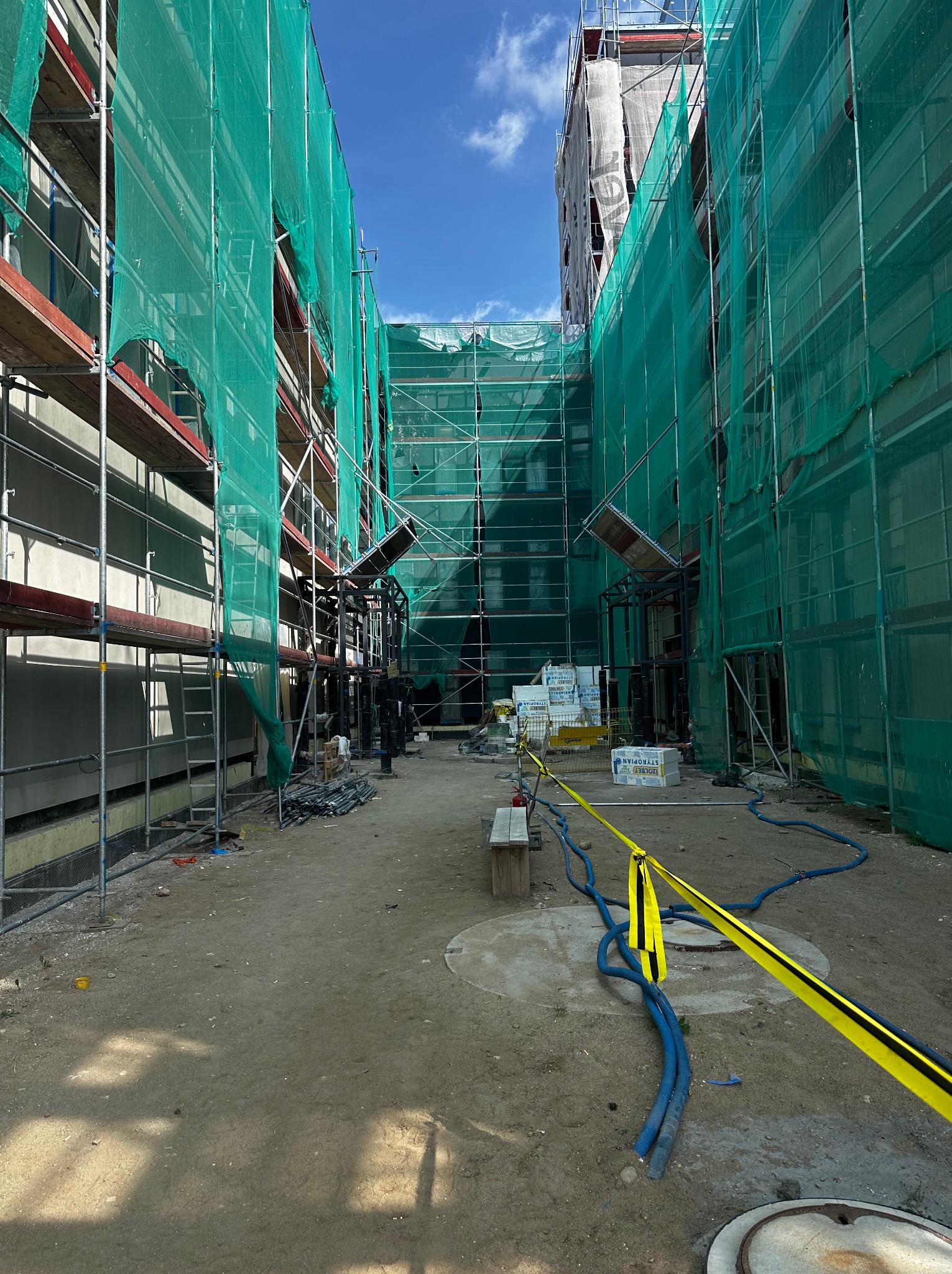 Budynek w rusztowaniach, osłonięty zieloną siatką. Widoczne materiały budowlane, zabezpieczenia i fragment nieba. Ujęcie z dołu, podkreślające wysokość konstrukcji.