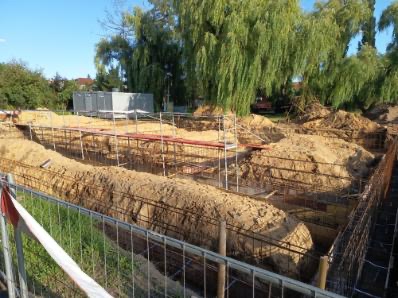 Wykopy pod fundamenty z widocznym zbrojeniem, przygotowane do zalania betonem. Prace ziemne w toku, widoczne rusztowania i zabezpieczenia.