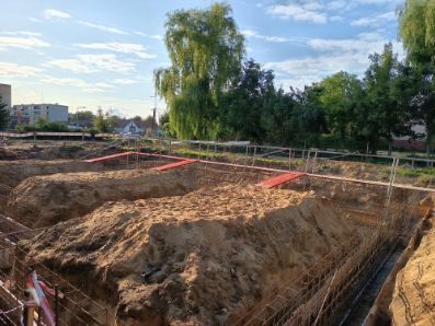 Wykopane fundamenty pod budowę domu, z widocznym zbrojeniem i deskowaniem. W tle budynki mieszkalne i drzewa, słoneczny dzień.