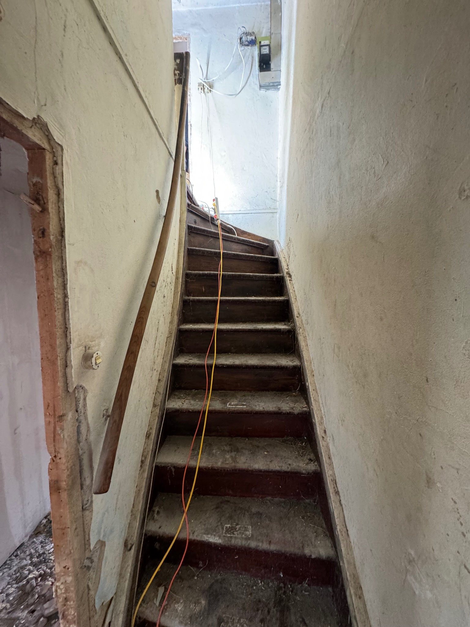 Klatka schodowa w trakcie remontu: widoczne stare, drewniane schody, ściany z odrapaną farbą i wystające przewody elektryczne. Ujęcie z dołu, podkreślające zakres prac.