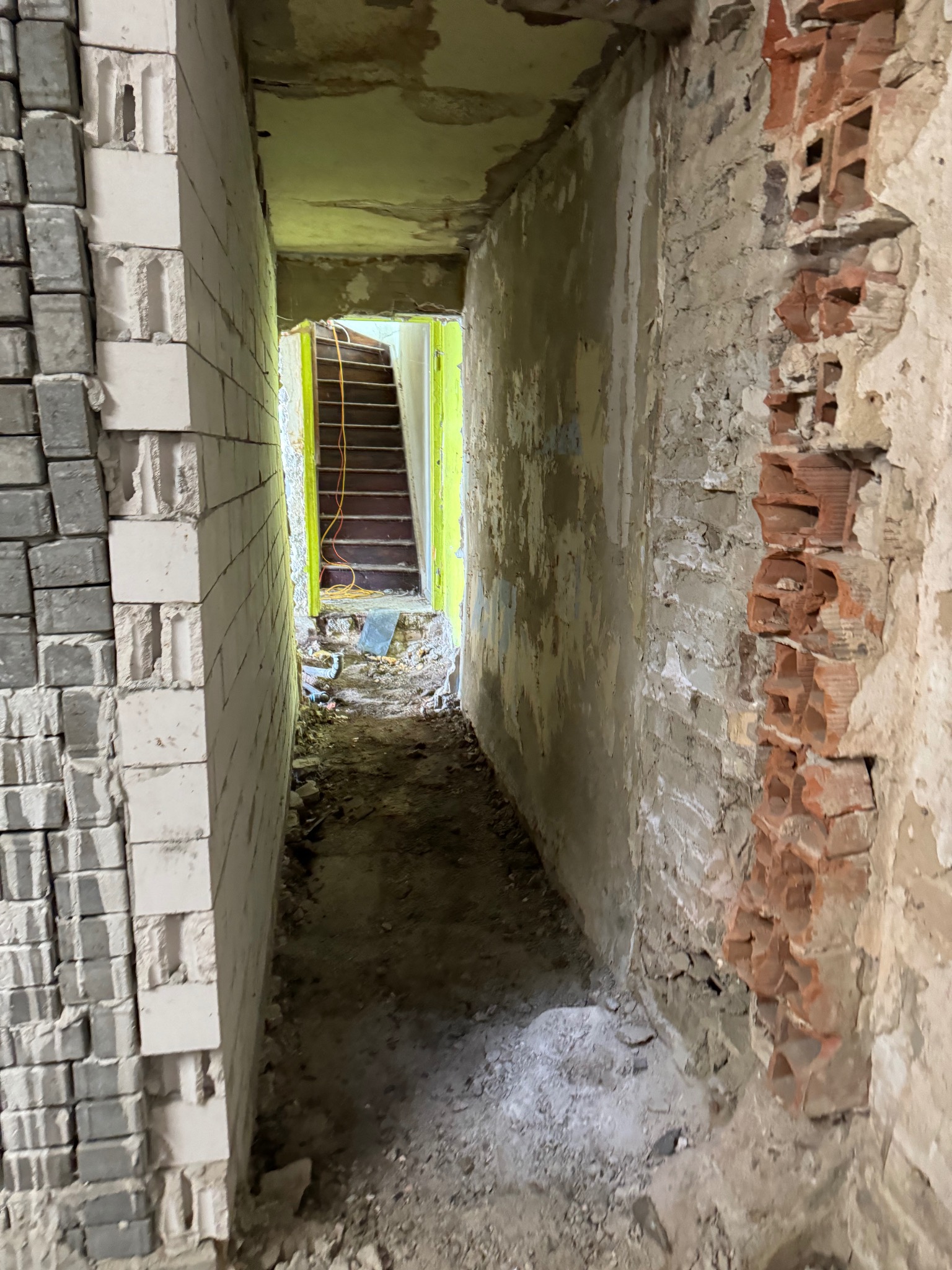 Wnętrze w trakcie remontu: widok na korytarz z odsłoniętą cegłą, stertą bloczków i schodami w tle, z elementami instalacji elektrycznej.