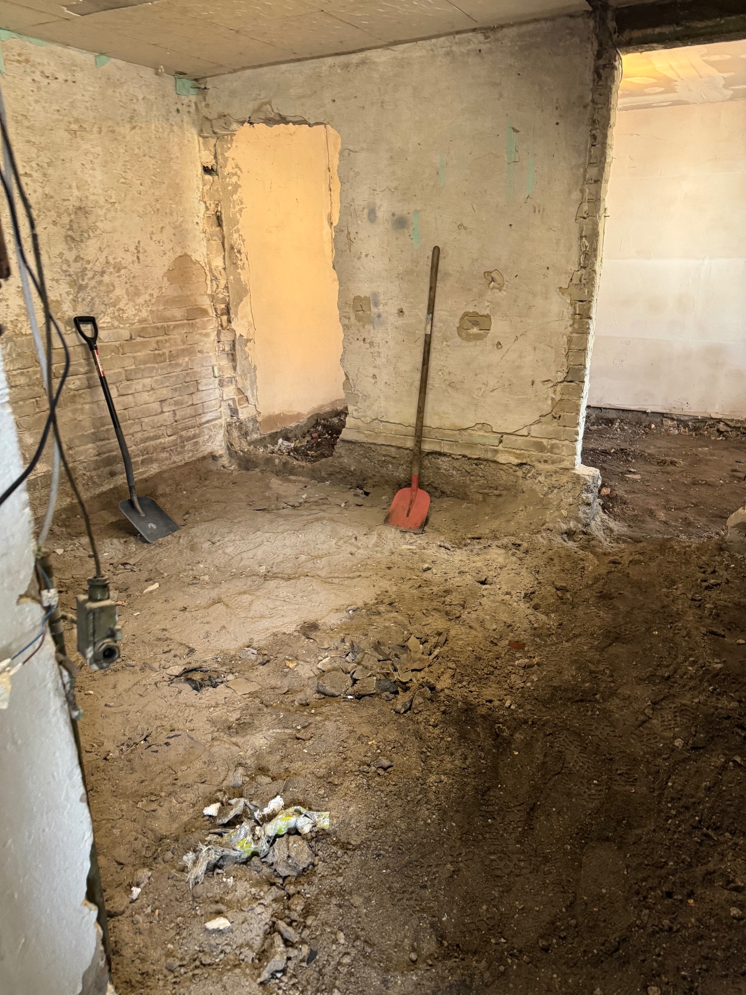 Wnętrze w trakcie prac przygotowawczych do remontu: odsłonięte cegły, resztki gruzu na podłodze, dwie łopaty opierają się o ściany. Widoczne ślady ingerencji w strukturę budynku.