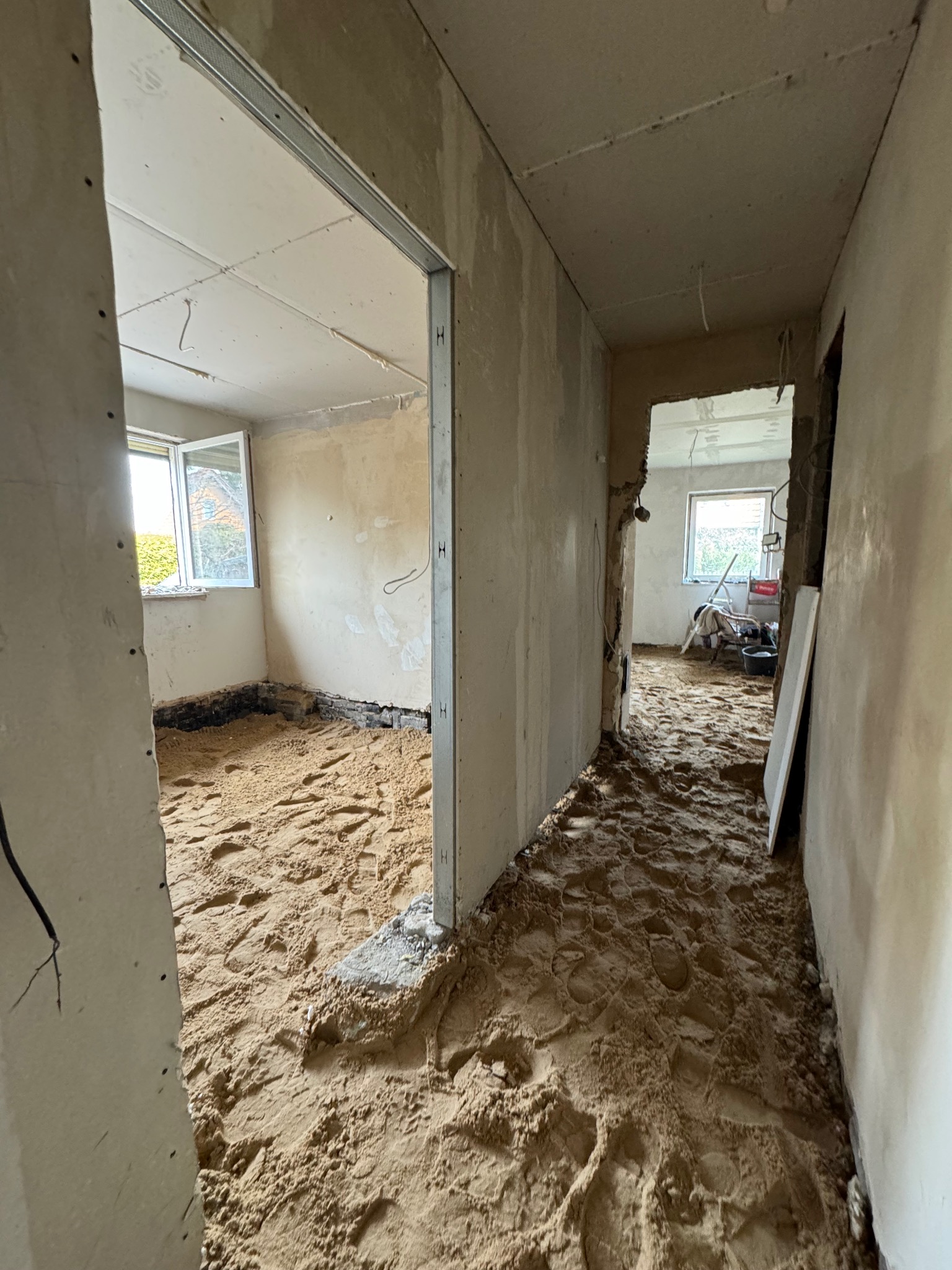 Wnętrze w trakcie remontu: piasek na podłodze, widoczne ślady stóp, metalowe profile ścianek działowych, okno z widokiem na zieleń. Prace przygotowawcze do wykończenia.