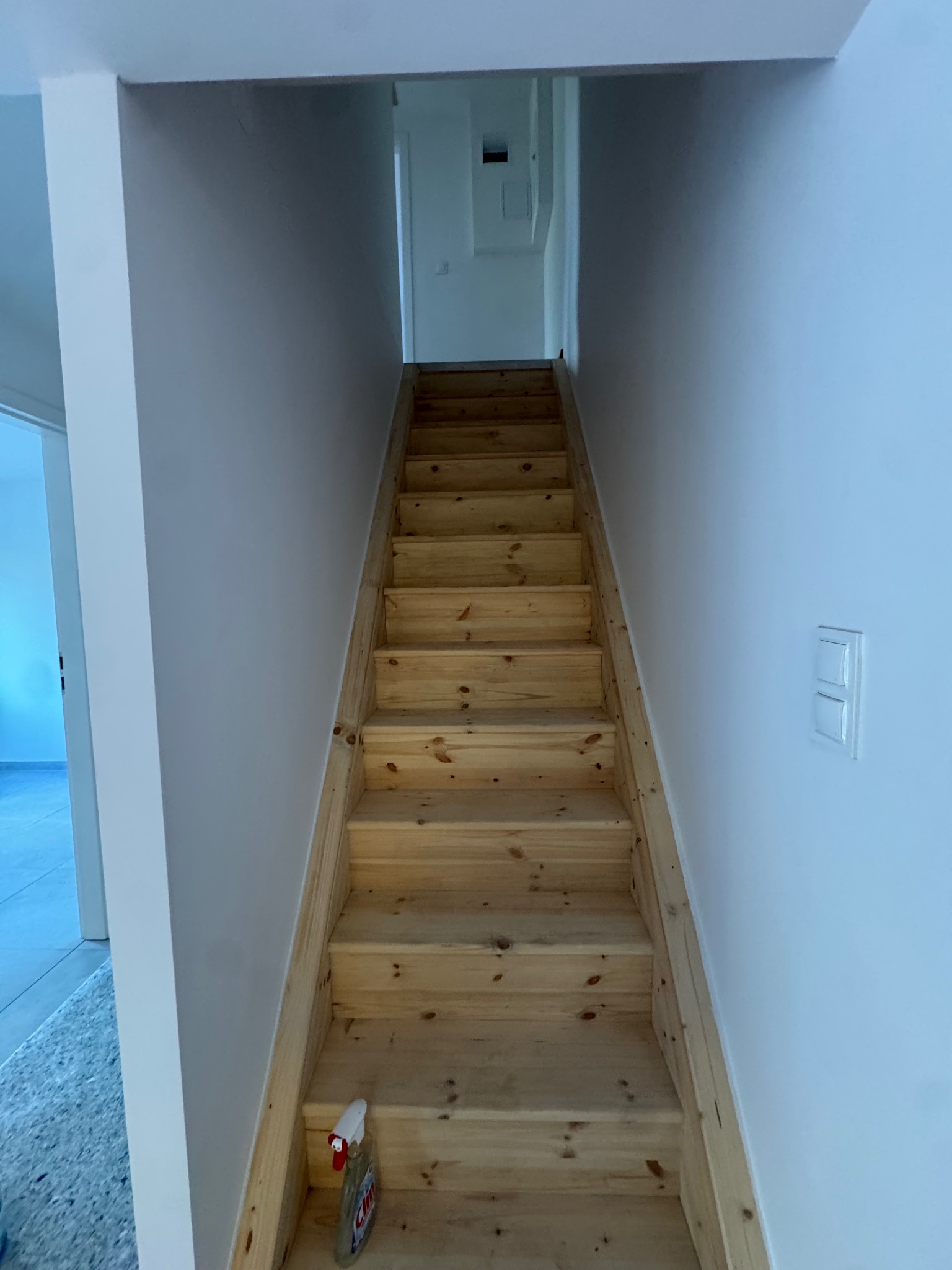 Drewniane schody prowadzące na górę, z butelką środka czyszczącego na dole. Białe ściany i widoczne gniazdko elektryczne. Ujęcie z dołu, perspektywa w górę.