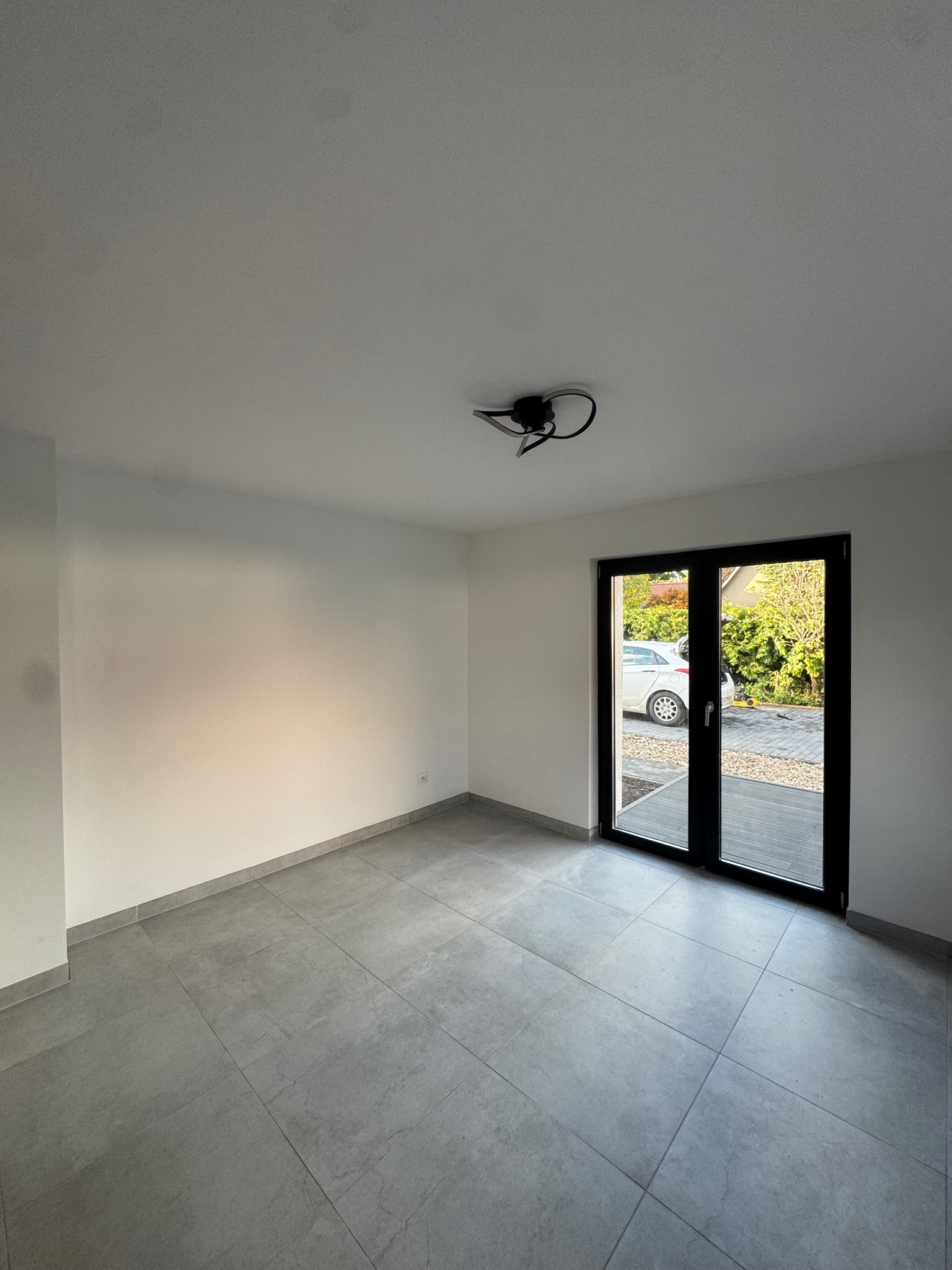 Jasne wnętrze z szarymi płytkami podłogowymi i czarnymi drzwiami balkonowymi. Widok na zewnątrz z samochodem i zielenią. Minimalistyczna lampa sufitowa.