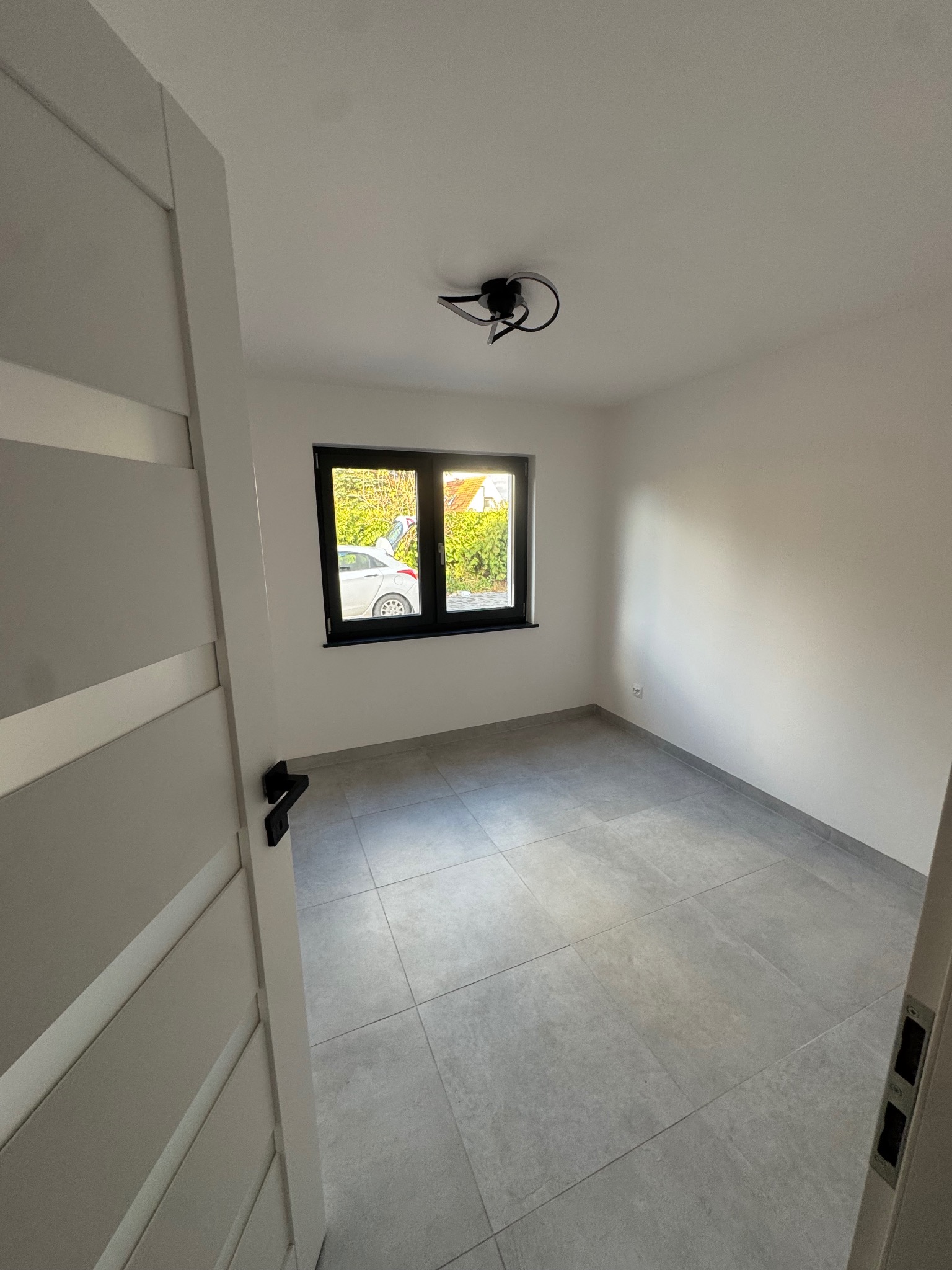 Pusty pokój z szarymi płytkami podłogowymi, białymi ścianami i czarną ramą okienną. Widok przez otwarte drzwi na fragment pokoju i okno z widokiem na zewnątrz.