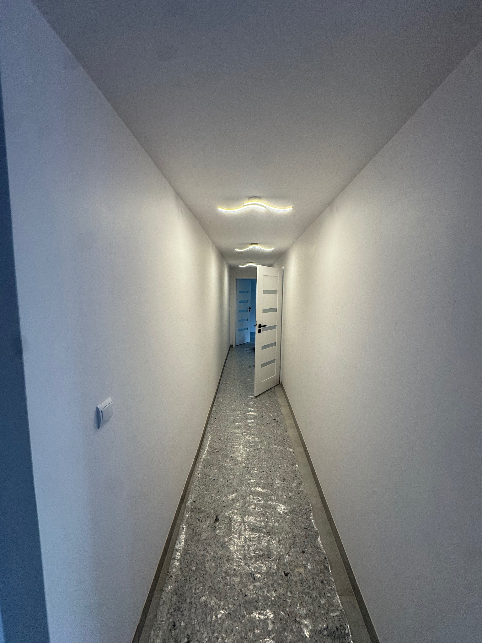 Biały korytarz z otwartymi drzwiami i nowoczesnym oświetleniem sufitowym. Podłoga zabezpieczona folią ochronną. Świeżo pomalowane ściany.